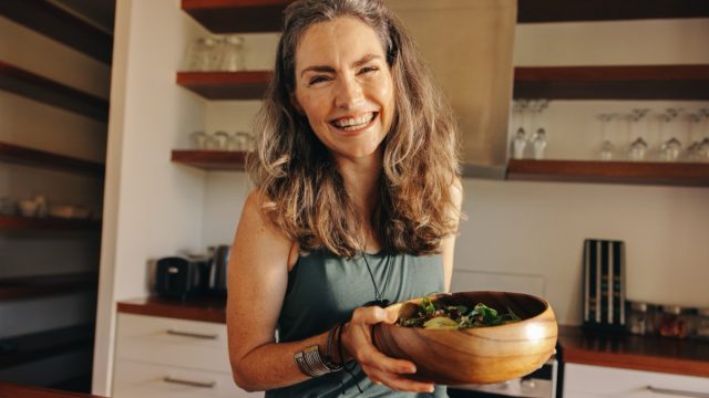Healthy senior woman smiling happily while holding a wholesome buddha bowl. Mature woman serving herself a delicious vegan meal at home. Woman taking care of her aging body with a plant-based diet.
