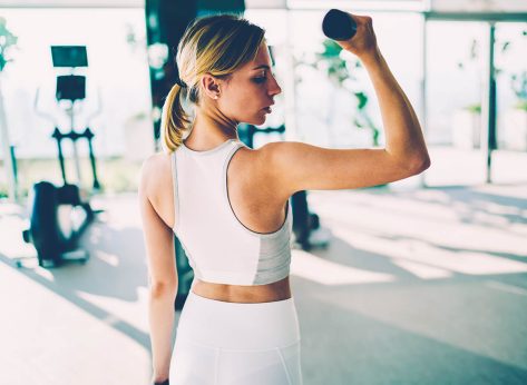 Young healthy woman dressed in white active clothing for training lifting weight dumbbells in modern gym interior. Athletic female person doing exercises fora biceps arms and shoulders indoors