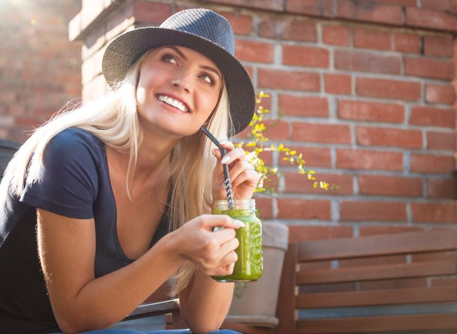 Blond young woman enjoying her green smoothie drink outside on terrace at sunset.