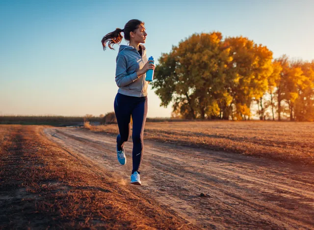 Woman running holding bottle of water in autumn field at sunset. Healthy lifestyle concept. Active sportive people