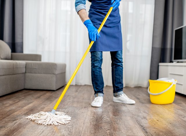 Man as a professional cleaner in blue uniform washing floor with mopping stick and bucket in the living room of the apartment