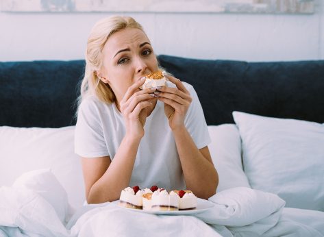 upset woman in pajamas looking at camera while eating cake in bed alone