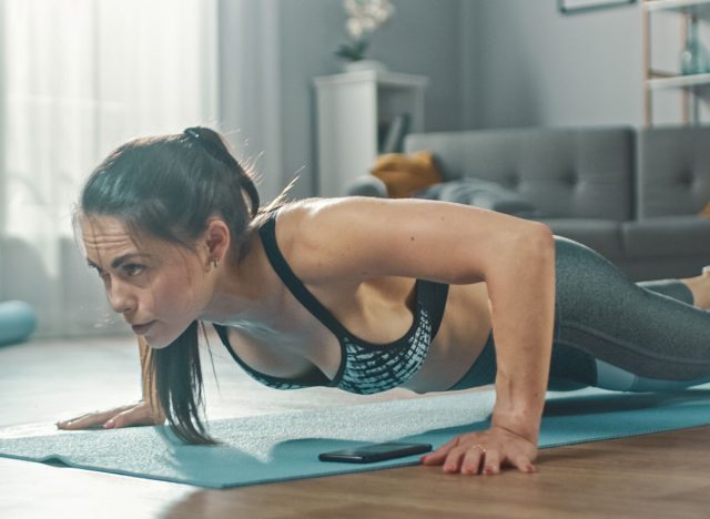 Close Up of a Beautiful Fitness Girl in an Athletic Top Doing Push Up Exercises While Using a Stopwatch on Her Phone. She is Training at Home in Her Living Room with Minimalistic Interior.
