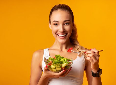 Sport And Diet. Cheerful Fitness Girl Eating Vegetable Salad Standing In Studio Over Yellow Background.