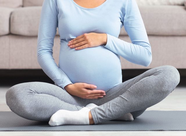 Close-Up Of Pregnant Woman Holding Her Belly, Sitting On Yoga Mat. Cropped image, panorama.