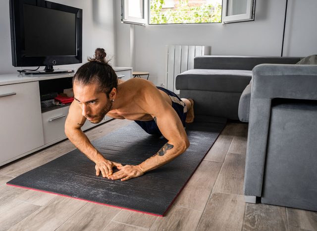 Young man in good physical shape, with bow tie, shirtless, and shorts, doing diamond-style push-ups on a mat on the floor in his bachelor's room, training