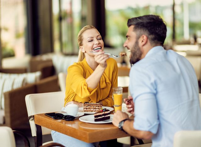 Happy woman having fun while feeding her boyfriend with a cake in a cafe.