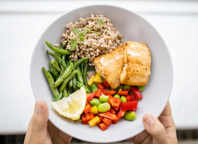 Hands holding salmon and buckwheat dish with green beans, broad beans, and tomato slices. Nutritious dish with vegetables and fish from above. Healthy balanced diet