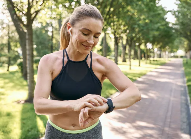 Morning jog. Beautiful sporty woman in sportswear and earphones looking at her watch, checking the result while running in a green park on a sunny summer day