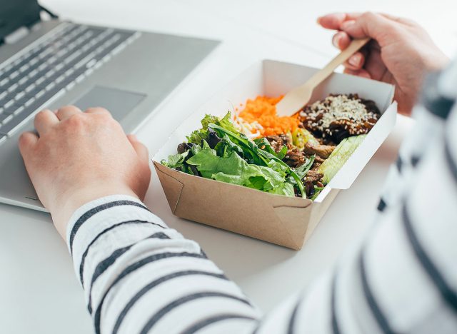 Woman having lunch from recycled bowl and using laptop. Concept of food delivery, quarantine, take out food