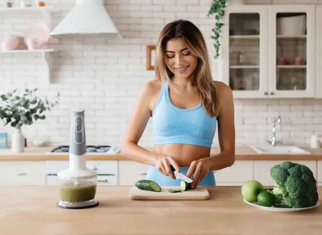 Waist-up photo of slim young woman smiling while cutting cucumber on wooden board