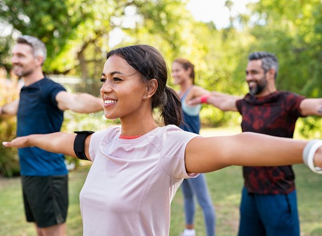 Group of multiethnic mature people stretching arms outdoor. Middle aged yoga class doing breathing exercise at park. Beautifil women and fit men doing breath exercise together with outstretched arms. 
