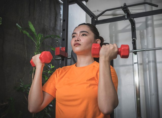 A serious and focused woman prepares to do a light set of arnold shoulder presses with vinyl plastic dumbbells at her home gym.