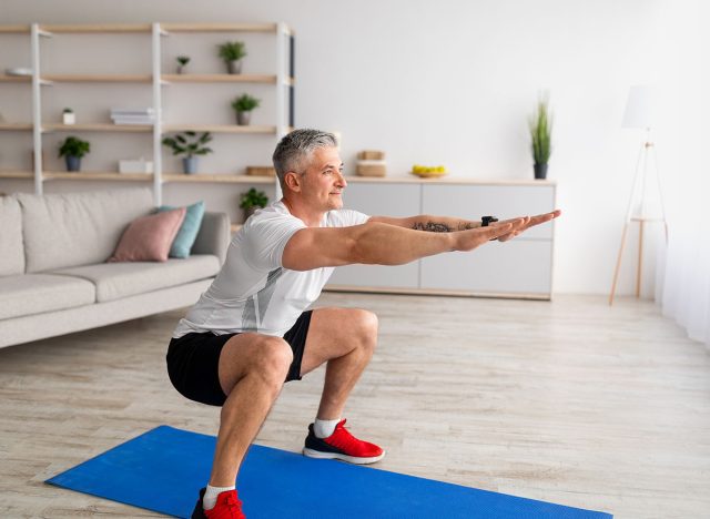 Stay home alone to workout. Fit mature man in sportswear doing squats in living room interior, exercising his legs on domestic training during coronavirus isolation, free space