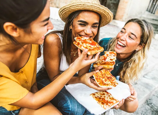 Three young female friends sitting outdoor and eating pizza - Happy women having fun enjoying a day out on city street - Happy lifestyle concept