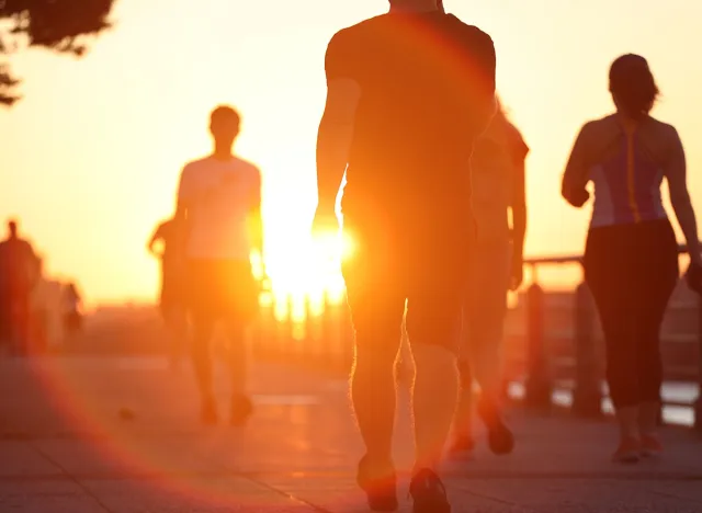 Group of unrecognized people exercise active walking on riverside boardwalk at sunset. Summer outdoor commuting fitness concept.