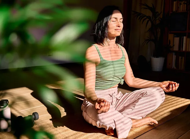 Woman practicing yoga and meditation at home sitting in lotus pose on yoga mat, relaxed with closed eyes. Mindful meditation concept. Wellbeing.