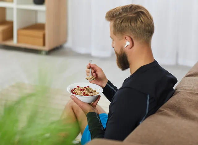 Healthy lifestyle. Athletic millennial man starts his day with healthy oatmeal with berries for breakfast. Unrecognizable man with wireless headphones in his ears sitting on sofa with bowl of oatmeal.