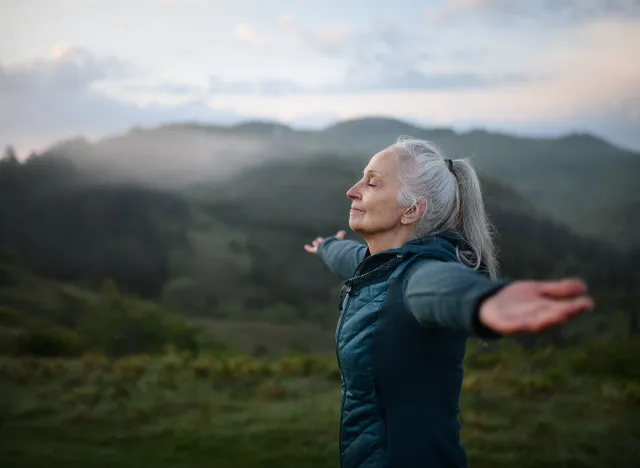 Senior woman doing breathing exercise in nature on early morning with fog and mountains in background.