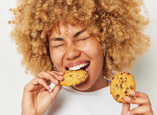 Close up shot of blonde curly haired woman eats cookies with chocolate bites deicious snack keeps eyes closed has sweet tooth dressed casually isolated over white background. Unhealthy eating