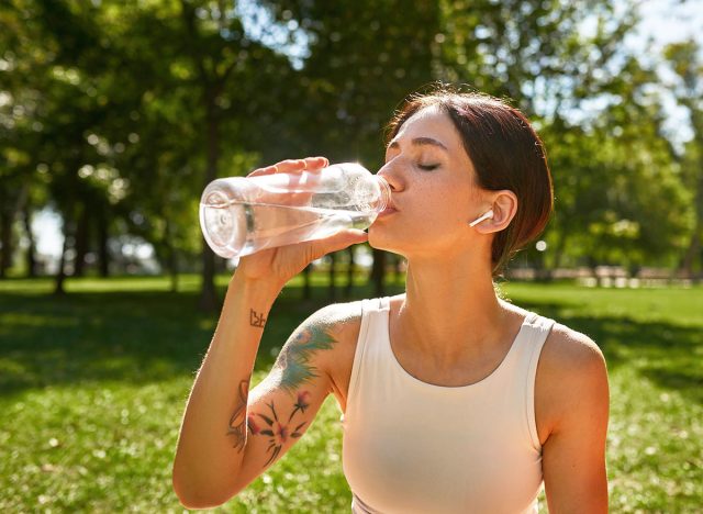 Cropped of young caucasian sportswoman drinking water after training on green lawn. Girl with tattoos and closed eyes wearing sportswear and earphones in sunny park. Healthy lifestyle