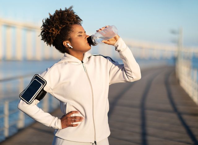 Young african american woman drinking water, having break during training outdoors, thirsty black female enjoying refreshing drink after jogging, wearing wireless earphones and armband, copy space