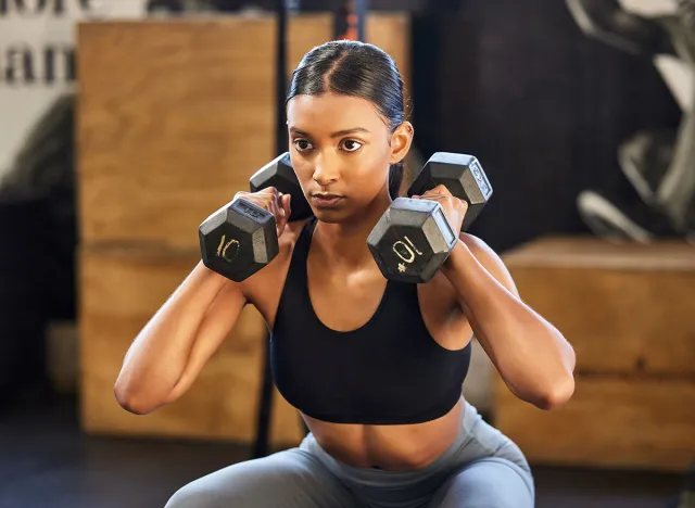 Fitness, squat or woman with dumbbells training, exercise or workout for powerful arms or muscles in gym. Dumbbell squats, bodybuilder or Indian girl athlete lifting weights or exercising biceps