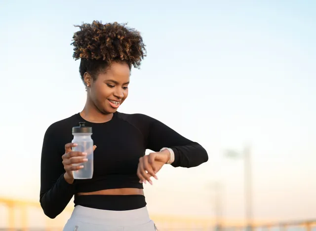 Smiling Black Female Jogger Checking Running Time On Smartwatch After Outdoor Training, Happy Young African American Woman Drinking Water And Checking Fitness Tracker Data On Watch, Copy Space