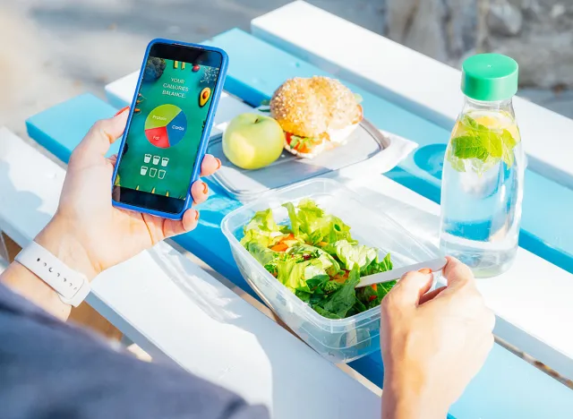 Close up woman using meal tracker app on phone while eating salad at picnic table in the park on a break. Healthy balanced diet lunch box. Healthy diet plan for weight loss. Selective focus