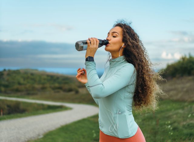 A girl is drinking water from her transparent bottle, a smart watch on her wrist, and headphones in her ears.