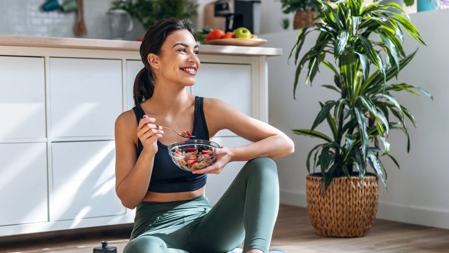Shot of athletic woman eating a healthy bowl of muesli with fruit sitting on floor in the kitchen at home