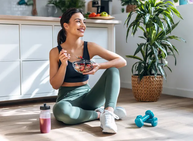 Shot of athletic woman eating a healthy bowl of muesli with fruit sitting on floor in the kitchen at home