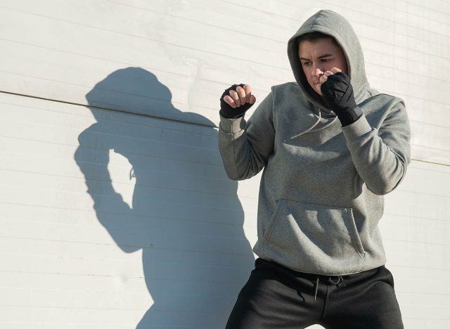 A man in a hoody trains boxing against a gray wall.