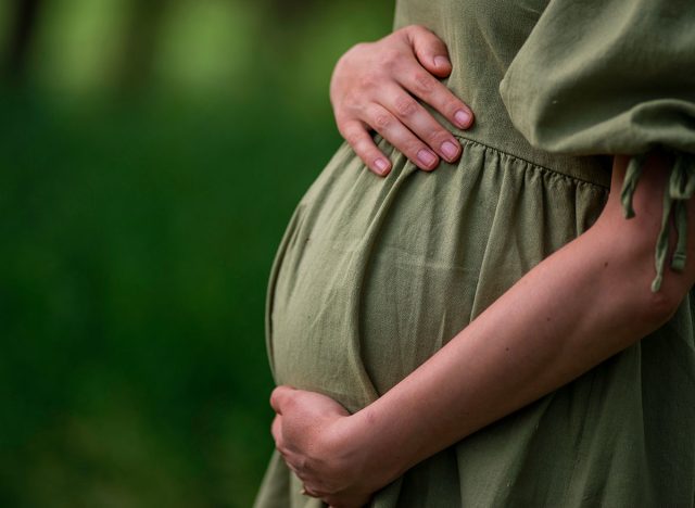 Caucasian Young Pregnant woman Tummy belly summer green dress touching stomach field expecting a baby relaxing outside nature park lake rural Beautiful magic 8 months enjoying life glasses hat