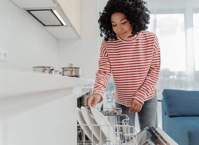 Portrait of beautiful African American woman wearing casual clothes loading dishwasher with dishes in kitchen at home. Housework concept