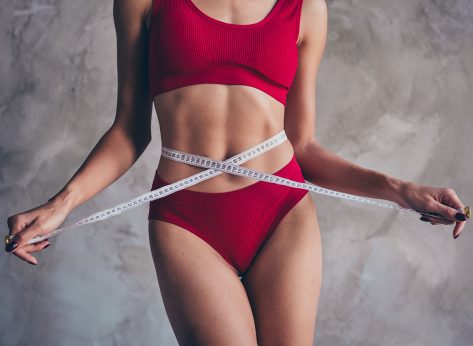 No retouch photo of body positive slim woman dressed red underwear keep measuring tape on waist isolated on gray concrete background