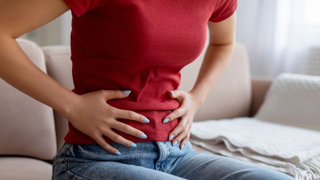 Unrecognizable woman experiencing abdominal pain at home, young female touching her stomach while sitting on couch in living room, feeling discomfort in digestion, cropped shot, closeup