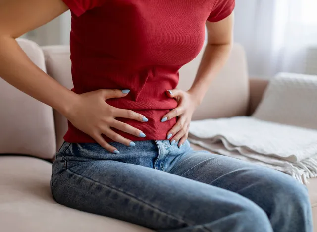 Unrecognizable woman experiencing abdominal pain at home, young female touching her stomach while sitting on couch in living room, feeling discomfort in digestion, cropped shot, closeup