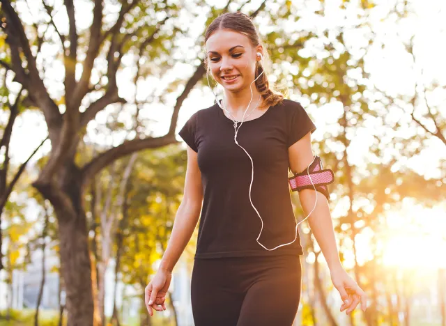Attractive young sporty smiling woman in sportswear walks in park at sunset with armband and earphones listening to music during training