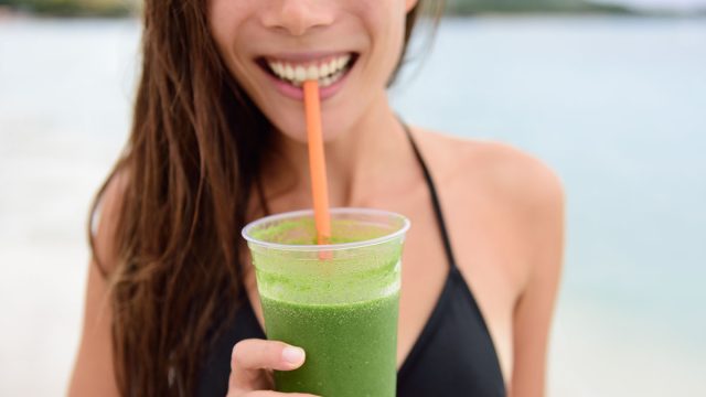Green smoothie. Woman holding green vegetable detox juice outside in bikini in summer sun on beach. Healthy lifestyle with beautiful mixed race Asian Caucasian female model taking a cleanse diet.