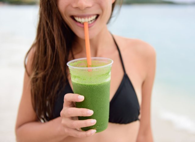 Green smoothie. Woman holding green vegetable detox juice outside in bikini in summer sun on beach. Healthy lifestyle with beautiful mixed race Asian Caucasian female model taking a cleanse diet.