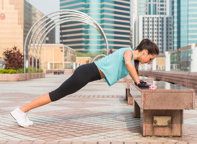 Fitness woman doing feet elevated push-ups on a bench in the city. Sporty girl exercising outdoors