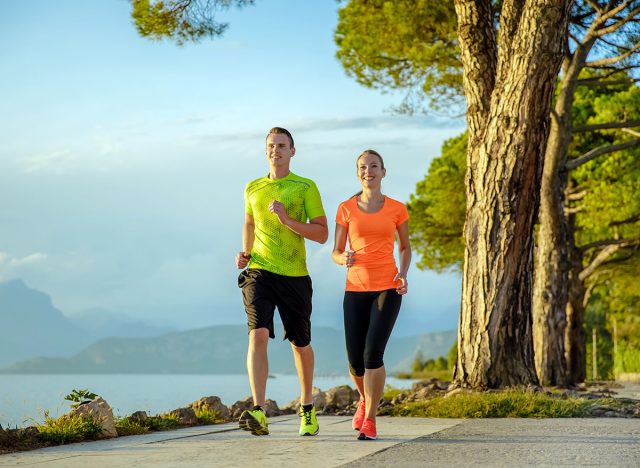 Young sexy couple is running along the promenade. they are doing their sport workout in the beautiful sundown along the beach. colorful dress, trees, water, mountains and a amazing blue sky.