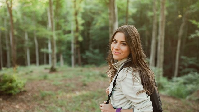 Young woman smiling at camera with backpack in the forest on sunset light in the autumn season, looking up, exploring the nature.