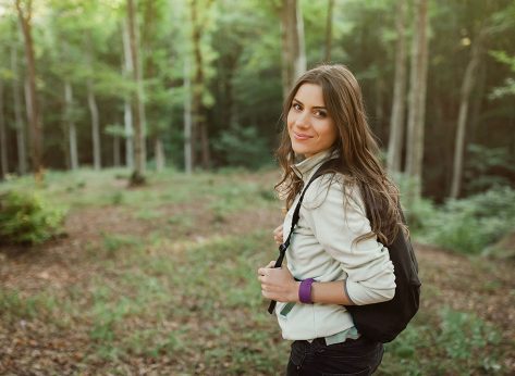 Young woman smiling at camera with backpack in the forest on sunset light in the autumn season, looking up, exploring the nature.