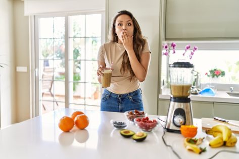 Young brunette woman drinking glass of smoothie at the kitchen covering mouth with hand, shocked and afraid for mistake.