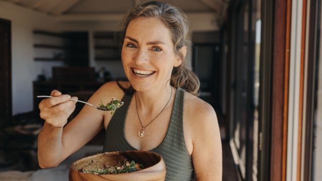 Happy vegan woman smiling at the camera while eating a vegetable salad from a bowl. Senior woman enjoying a plant-based breakfast after a home workout. Mature woman taking care of her ageing body.
