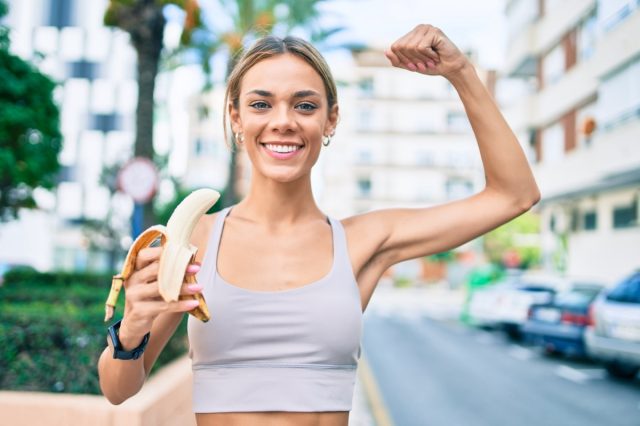 Young cauciasian fitness woman wearing sport clothes training outdoors eating healthy banana and showing proud arm muscle.