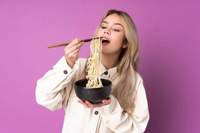 Woman isolated on purple background holding a bowl of noodles with chopsticks and eating it.