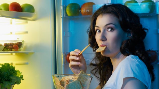 Woman eating snacks in night next to the opened fridge.